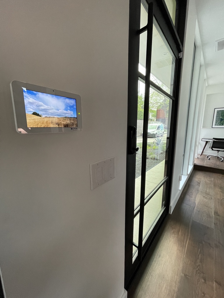 Wall-mounted security control panel with a built-in camera beside a glass door fitted with a smart lock