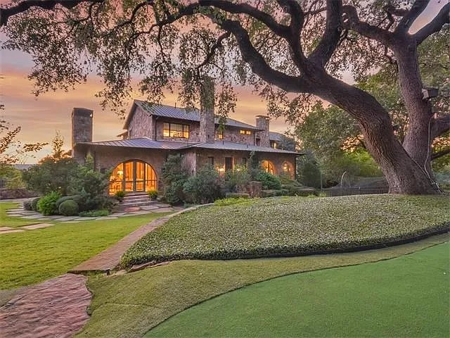 Stone residence exterior at dusk beneath a sprawling live oak with illuminated windows and grounds